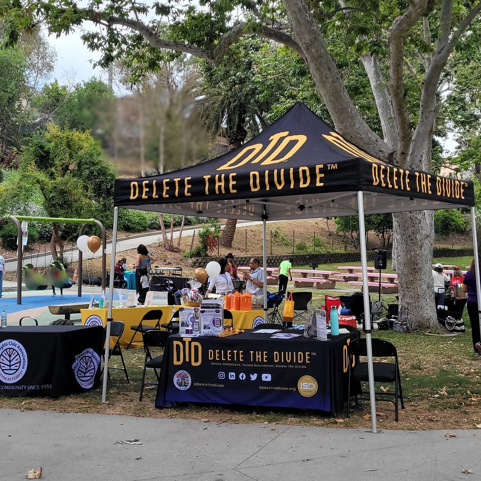 A park event with "Delete The Divide" tents, people, and tables near a playground.