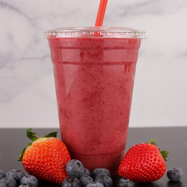 Berry smoothie in a clear cup with a red straw, surrounded by fresh strawberries and blueberries.