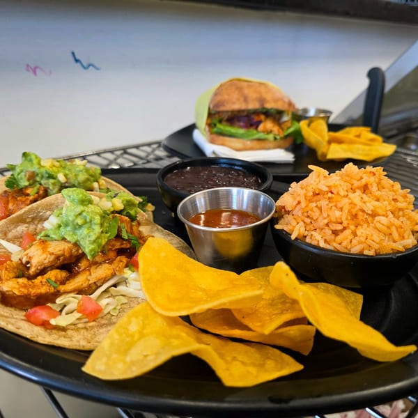 Two chicken tacos with guacamole, rice, black beans, chips, and a burger on a black tray.