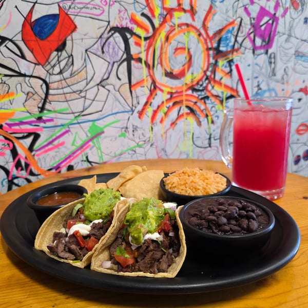 Two tacos with guacamole, rice, beans, chips, salsa, and a red drink on a wooden table.